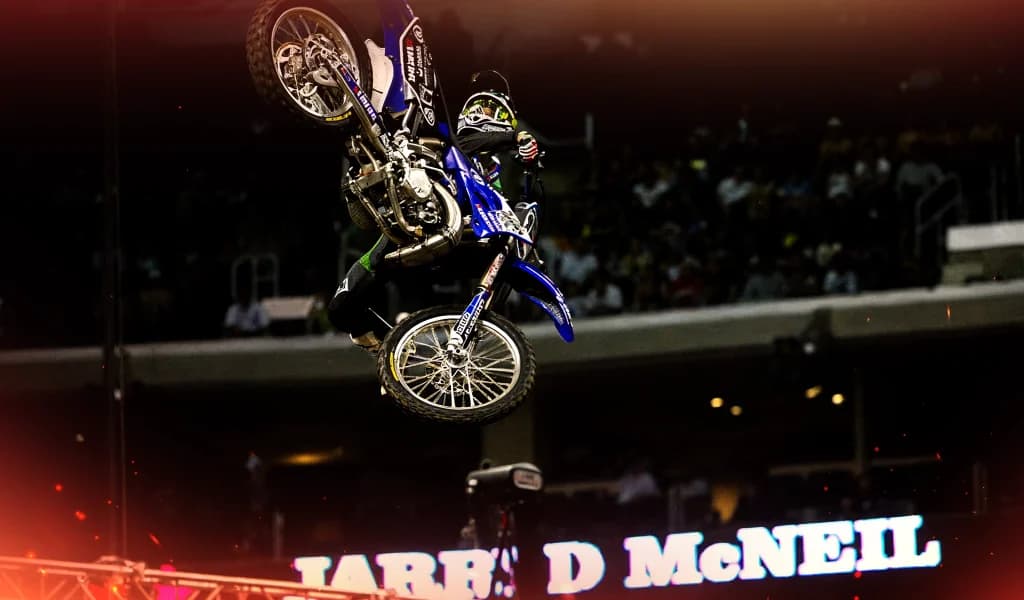 A motocross rider, Jarryd McNeil, performing a high aerial trick above a stadium crowd during a night competition.