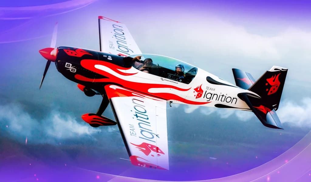 Red, white, and black aerobic airplane wrapped in Team Ignition branding flying at a steep angle through a cloudy sky.