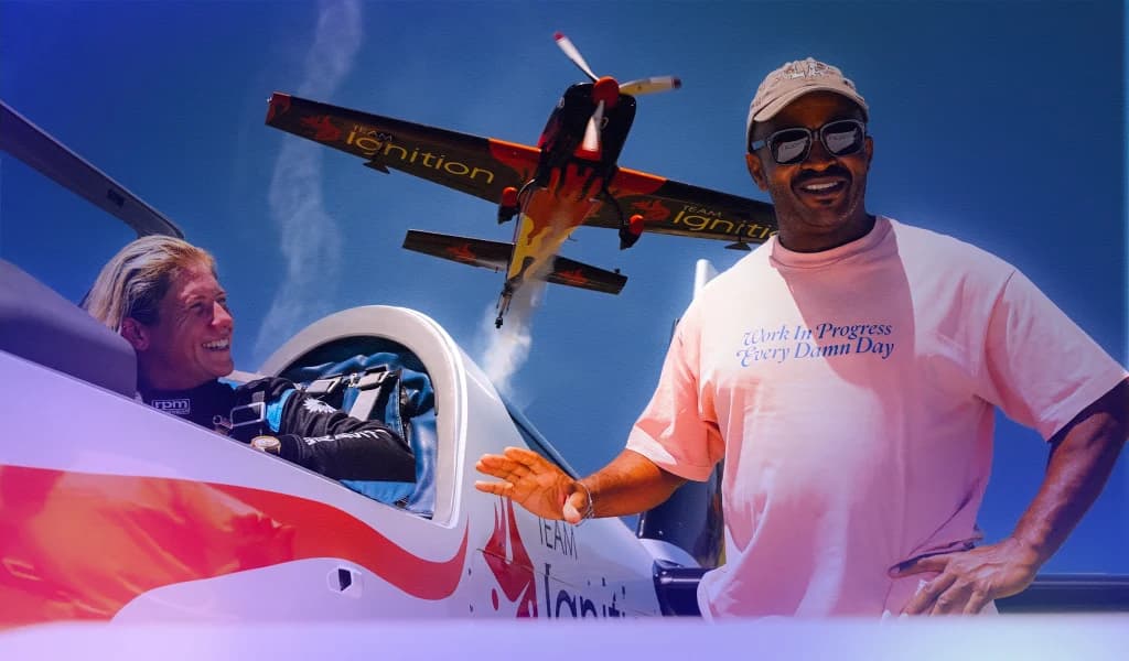 Aerobic pilot Emma McDonald seated in a cockpit while another person stands beside the aircraft, with a second plane flying overhead.