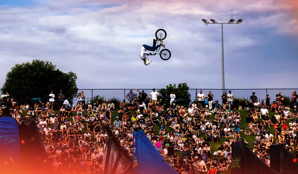 BMX rider performs a backflip mid-air above a large outdoor crowd.