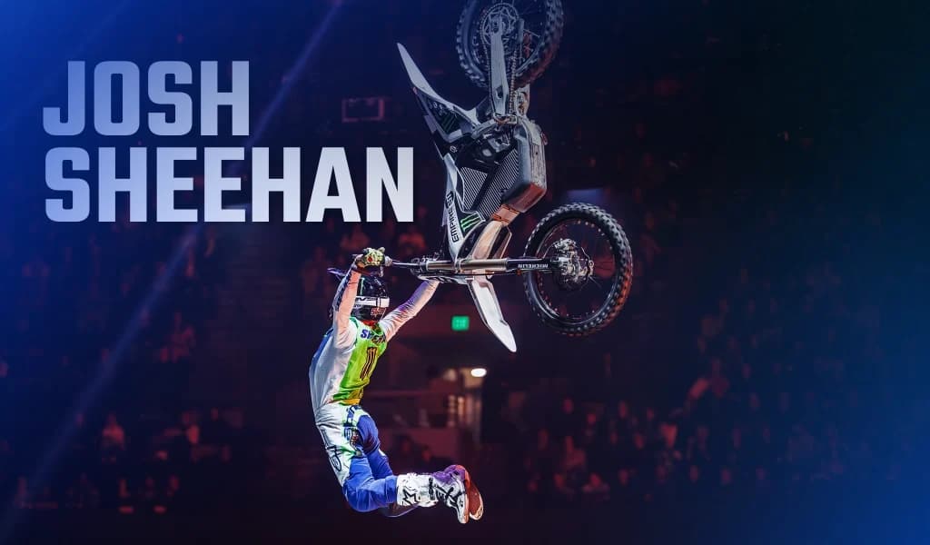 A motorbike athlete hanging from the bike during a high aerial above a crowded arena.