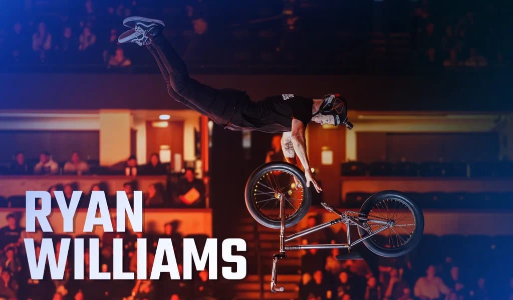 A BMX rider stretched out horizontally while holding the bike during an aerial trick in an arena.