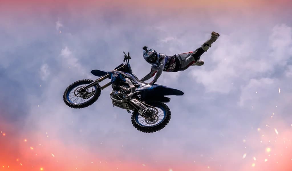 A motocross rider extends their body behind the bike while performing an aerial trick against a cloudy sky.