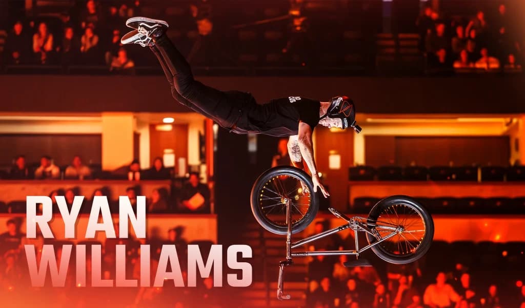 BMX rider Ryan Williams extending into a horizontal position while holding the handlebars during an indoor arena performance.