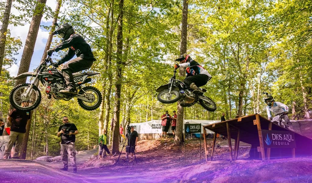 Two pit bike riders at Pastranaland midair over a dirt jump with additional riders and cameras positioned around the course.