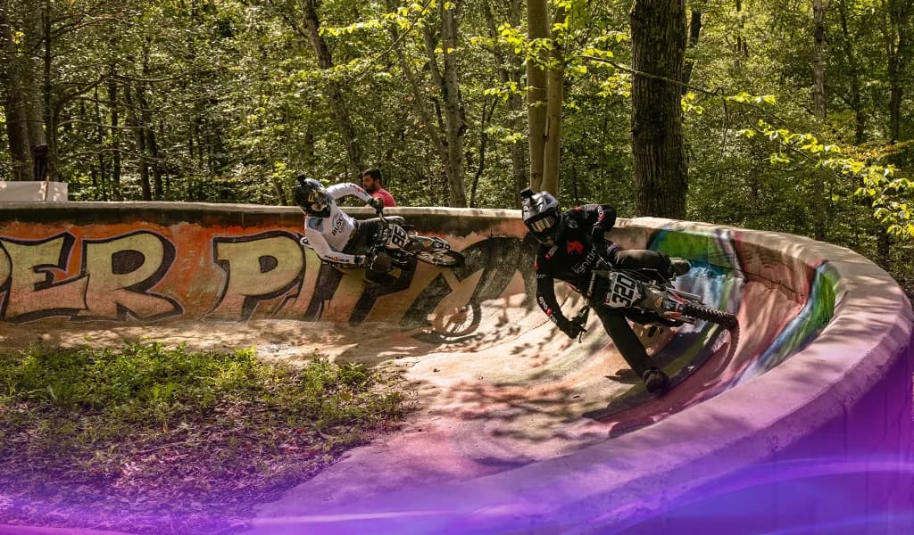 Two pit bike riders leaning into a curved concrete berm on a wooded track covered in shadows.