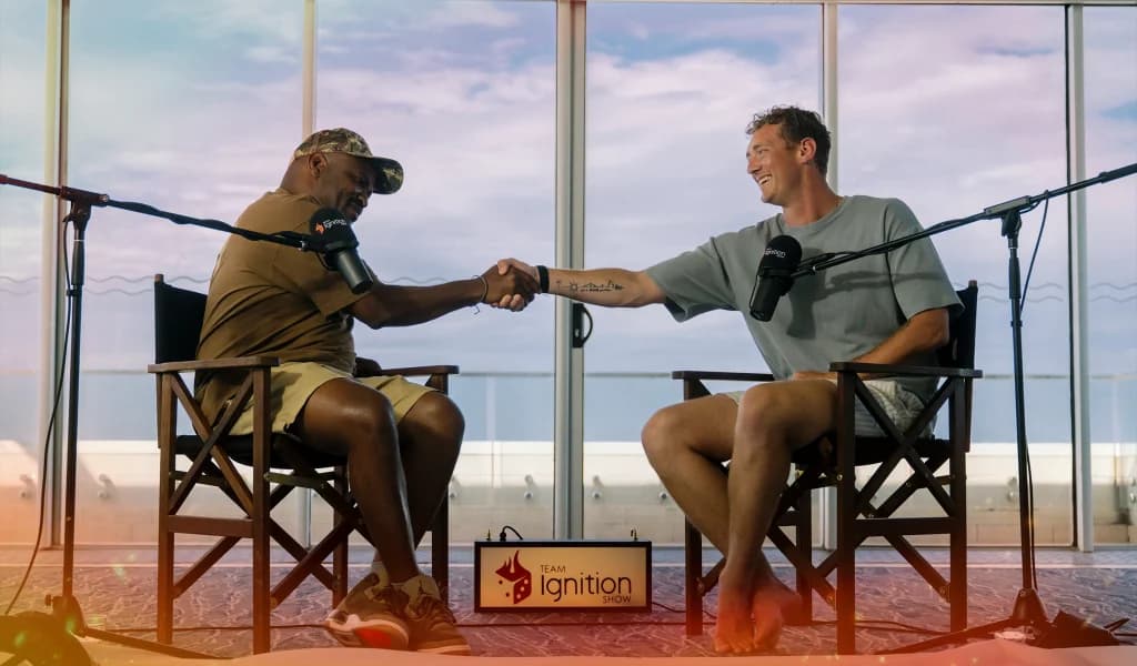 Two people seated in chairs shaking hands during a podcast-style interview with microphones on stands.