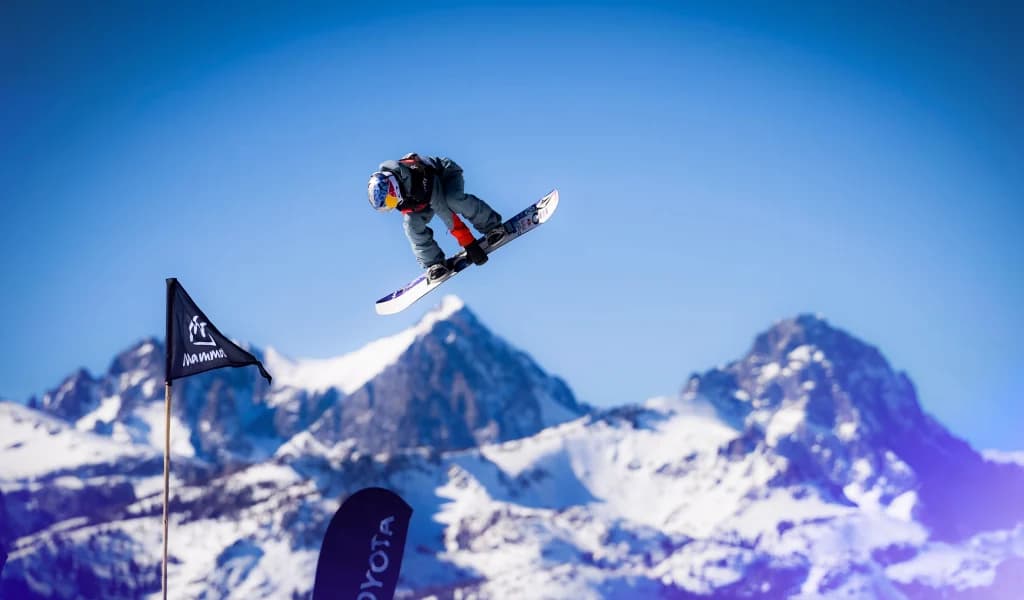 Snowboarder airborne over a jump with snow-covered mountains in the background.