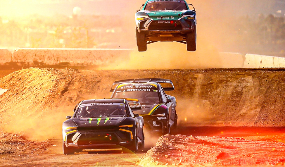 Three Nitrocross racing cars on a dirt track, one airborne in a jump